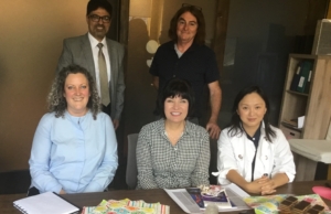 Photograph of three people seated at a table and two standing behind them smiling for their photo being taken